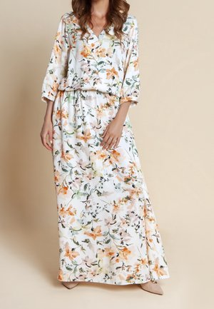 Frau trägt ein langes weißes Kleid mit orange- und grünem Blumenmuster, Dreiviertelärmeln und beigen spitzen Schuhen.