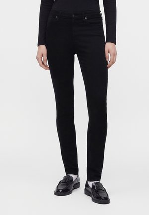 Personne portant un jean noir slim, un haut noir à manches longues, des chaussettes blanches et des mocassins en cuir noir, debout devant un fond clair uni.