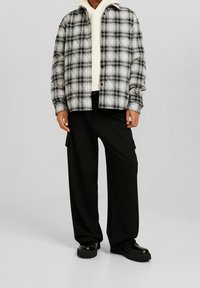 Svart och vit rutmönstrad skjorta med knapppdesign, bärs över en krämfärgad hoodie, kombinerat med lösa svarta cargobyxor och svarta skor.