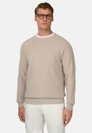 Homme aux cheveux courts et lunettes portant un pull beige texturé et un pantalon blanc, debout devant un fond clair uni.