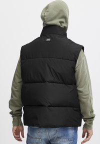 Schwarze gepolsterte Weste mit hohem Kragen, die über einem hellgrünen Langarmshirt getragen wird. Sie verfügt über einen strukturierten Stoff und genähte Details.