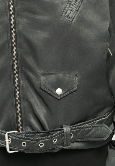 Svart matt jacka med en diagonalt placerad dragkedja, ficka med tryckknapp och ett justerbart bälte med metall detaljer. Texturerat material förstärker detaljerna.