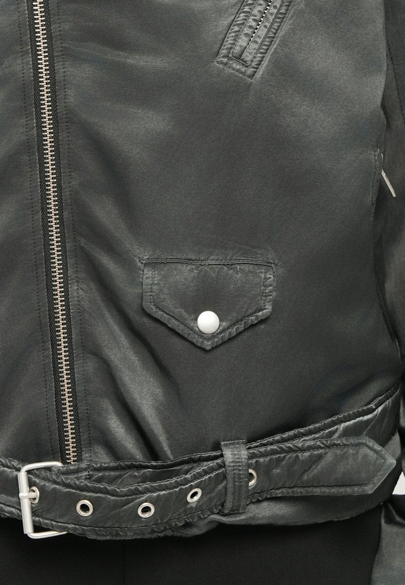Black matte jacket featuring a diagonal zipper, snap button pocket, and an adjustable belt with metal hardware. Textured material enhances detail.