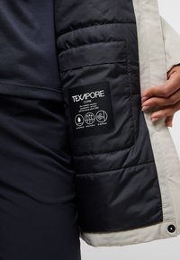Veste légère beige avec une doublure intérieure matelassée sombre. Présente une étiquette Texapore avec des icônes imperméables, respirantes et coupe-vent.