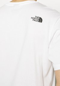 Person som bär en vit T-shirt med en liten svart "The North Face"-logotyp på den övre delen av ryggen nära axeln.
