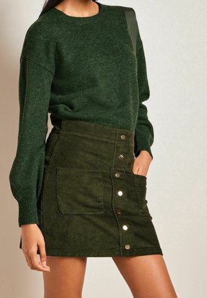 Femme portant un pull en maille vert foncé et une jupe en velours côtelé verte taille haute, boutonnée sur le devant avec des poches.