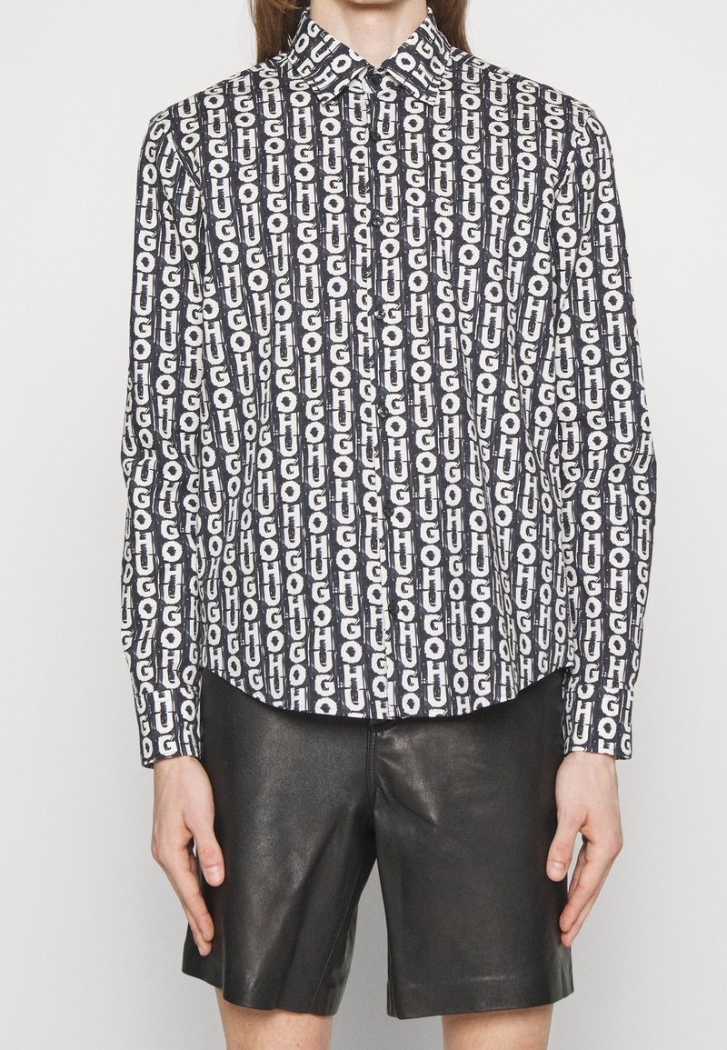 Personne portant une chemise noire et blanche à manches longues avec un texte répétitif vertical et un short en cuir noir, debout devant un fond uni.