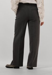 Grijze, op maat gemaakte broeken met een recht toe recht aan ontwerp, voorzien van riemlussen, achterzakken en een subtiele textuur. Gedragen met een witte, geborduurde blouse.