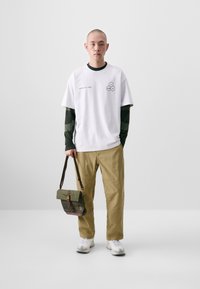 White oversized t-shirt with black text and hexagonal design, layered over a dark green long-sleeve shirt. Khaki pants and a green messenger bag.
