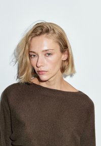 Donna bionda con capelli corti indossa un maglione marrone largo, in piedi davanti a uno sfondo grigio chiaro uniforme, guardando leggermente di lato.