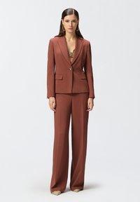 Traje color óxido que presenta una chaqueta ajustada con solapas y dos bolsillos frontales, combinada con pantalones de pierna ancha. Tejido suave con un corte estructurado.