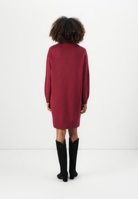 Knie-langes, bordeauxrotes Strickkleid mit hohem Kragen und lockerer Passform. Getragen mit schwarzen kniehohen Stiefeln, die eine spitze Zehenpartie und einen leichten Absatz haben.