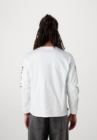 Camicia bianca a maniche lunghe con scollo rotondo e testo grafico nero sulla manica destra. Il tessuto appare liscio; il retro è semplice.