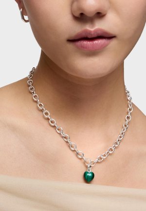 Srebrny naszyjnik z łańcuszkiem o okrągłych ogniwach i małą zieloną zawieszką w kształcie serca noszony na gołej szyi i ramionach.