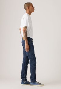 Λευκό κοντομάνικο μπλουζάκι, μπλε τζιν τζιν και μπλε αθλητικά παπούτσια. Ορατά τατουάζ στο μπράτσο. Πλευρική όψη σε φωτεινό φόντο.