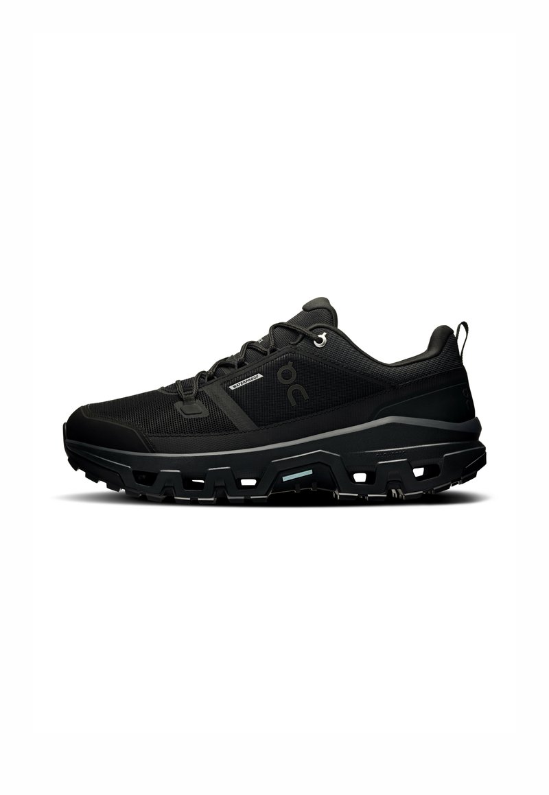 Chaussures de sport noires avec tige en mesh, détails texturés, semelle prononcée avec des reliefs profonds, et accents réfléchissants sur un fond blanc.