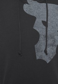 Gros plan sur un sweat à capuche noir avec un motif graphique gris foncé représentant une tête de taureau sur la poitrine et des cordons noirs suspendus à l’avant.