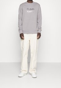 Felpa grigia con logo "Calvin Klein" bianco, collo rotondo, polsini a costine, abbinata a pantaloni beige chiaro a gamba larga e sneakers bianche.