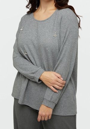 Femme portant un haut gris à manches longues orné de petites décorations en cristal, tenant un bras croisé sur sa taille.