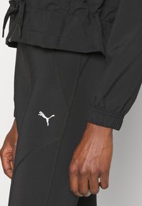 Zwarte sportjas met elastische boorden en een verstelbare taille, gecombineerd met zwarte leggings met een reflecterend Puma-logo en gemêleerde naden.
