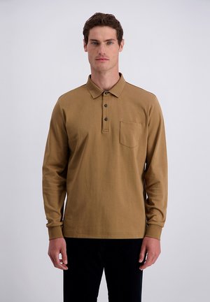 Uomo che indossa una polo marrone chiaro a maniche lunghe con taschino sul petto e pantaloni neri, in piedi davanti a uno sfondo chiaro uniforme.