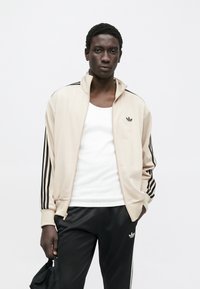 Svetlosmeđa jakna na patentni zatvarač s crnim dizajnom tri pruge na rukavima. Nošena preko bijelog topa. Crne sportske hlače s detaljem logotipa.