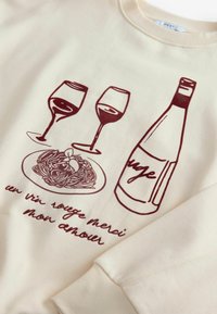 Sweatshirt crème avec un graphique maroon représentant deux verres de vin, une bouteille et une assiette de spaghetti, accompagné de texte stylisé.