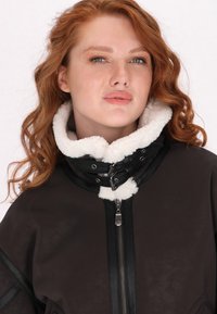 Ruskea tekonahkainen takki valkoisella fleece-kauluksella, vetoketjulla ja mustilla yksityiskohdilla. Ominaisuuksiin kuuluu rento istuvuus ja karheapintainen pinta.