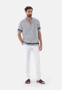 Camicia a maniche corte bianca con un motivo geometrico blu navy, abbinata a pantaloni bianchi e sandali neri, caratterizzata da una vestibilità rilassata e colletto.