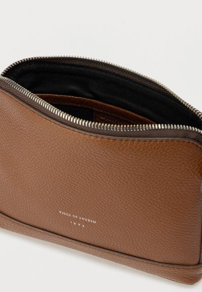 Καφέ δερμάτινο πουγκί με υφή, ασημί φερμουάρ μερικώς ανοιχτό, μαύρο υφασμάτινο εσωτερικό και το λογότυπο "Tiger of Sweden 1903" τυπωμένο στην πρόσοψη.