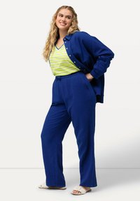 Tvådelad outfit bestående av en blå texturerad skjorta med knappar och matchande löst sittande byxor, tillsammans med en grön och vit randig tanktopp och sandaler.