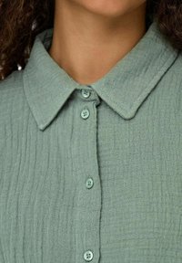 Camicia di un verde chiaro, texture con colletto e quattro bottoni abbinati sul davanti. Il tessuto ha un aspetto stropicciato.