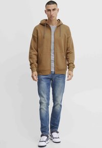 Sweat-shirt à capuche marron avec zip, poches avant et cordons de serrage contrastants sur un shirt gris. Associe avec un jean bleu et des baskets blanches.