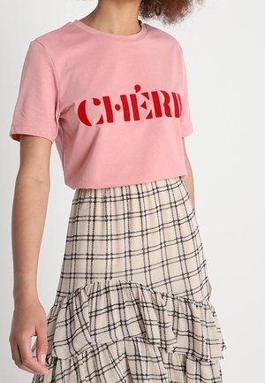 Pink cotton crop t-shirt with red block lettering "CHÉRI” paired with a beige, plaid, tiered skirt featuring black lines and ruffles.