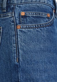 Pantalones vaqueros de mezclilla azul oscuro con remaches de cobre, costuras naranjas y textura suave. El diseño incluye un bolsillo delantero y detalle en la cintura.