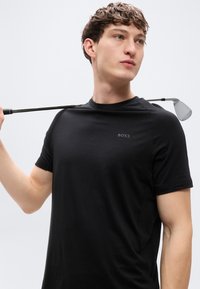 Μαύρο κοντομάνικο T-shirt με στενή εφαρμογή, με διακριτικό λογότυπο "BOSS" στο στήθος. Ένα γκολφ μπαστούνι ακουμπά στον ώμο.