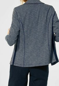 Blazer textured blu navy con un design aderente, caratterizzato da revers a punta e due spacco laterali. I dettagli in filo chiaro aggiungono un tocco di eleganza.