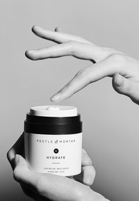 Pestle & Mortar Hydrate est un pot rond blanc avec un couvercle noir, arborant un design minimaliste et une étiquette textuelle. Une main se tend vers le couvercle.