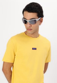 Tricou din bumbac galben cu un mic logo "GANT" în culorile albastru și roșu. Ochelari de soare gri, clar, cu lentile închise la culoare. Modelul are părul scurt și închis la culoare.