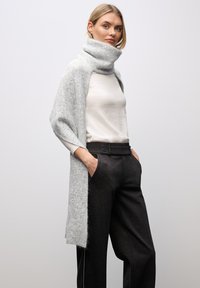 Sciarpa lavorata a maglia di colore grigio chiaro con colletto alto, indossata sopra una maglietta bianca e pantaloni neri. Texture morbida, frontale aperto. Design minimalista.