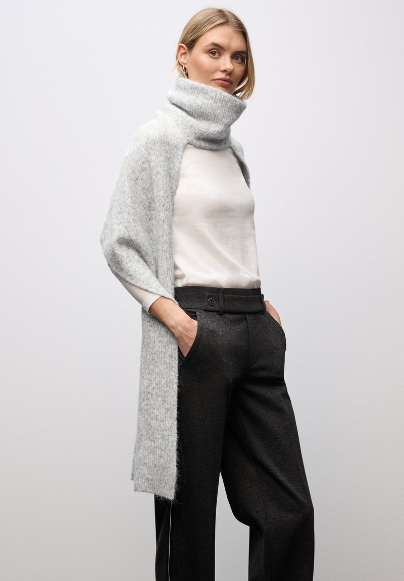 Sciarpa lavorata a maglia di colore grigio chiaro con colletto alto, indossata sopra una maglietta bianca e pantaloni neri. Texture morbida, frontale aperto. Design minimalista.