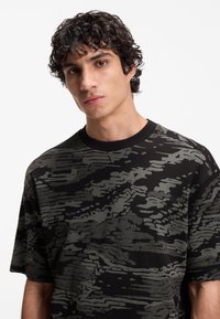 Νέος άνδρας με σγουρά σκουρόχρωμα μαλλιά, που φοράει μαύρο και γκρι πουκάμισο με σύντομο μανίκι και αφηρημένο σχέδιο, ουδέτερη έκφραση, λευκό φόντο.