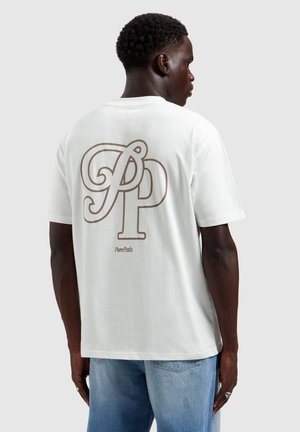 Man in wit T-shirt met grote beige letters "PP" en de tekst "Pure Path" op de rug, gecombineerd met lichtblauwe spijkerbroek, met de rug naar voren gekeerd.