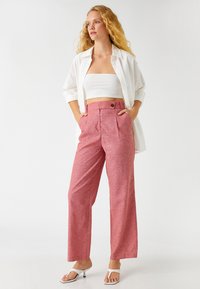 Pantalones de pierna ancha, de color rojo claro, confeccionados en una tela texturizada, con plisados frontales y cierre de botón. Combinados con un top blanco corto y una camisa.