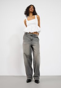 Fehér crop top fodros aljjal, amelyet magas derekú, világosszürke farmer széles szárú nadrággal párosítottak. A modellen fekete hegyes orrú magassarkú cipő van.