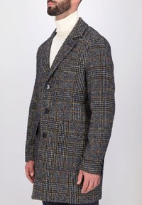 Cappotto di lana a quadri nelle tonalità di grigio, arancione e nero. Presenta un colletto a punta, chiusura a doppio bottone e tasche laterali. Texture liscia.