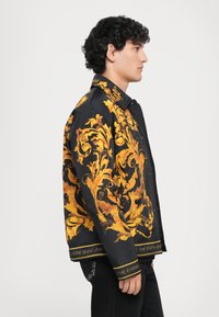 Tânăr îmbrăcat într-o jachetă neagră cu un model floral baroc strălucitor de culoare galbenă, stând de profil pe un fundal simplu deschis.