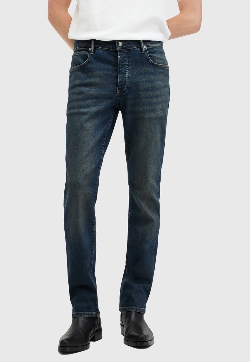 Jeans de mezclilla azul oscuro con un corte slim, diseño de cinco bolsillos y un leve desgaste. Cuenta con cierre de botón y cremallera. Textura suave.