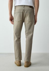 Person, der steht und beigefarbene Jeans mit geradem Bein, hellbraune Wildledersneaker und ein weißes Hemd trägt, von hinten vor einem schlichten Hintergrund gezeigt.