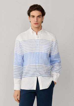 Jeune homme portant une chemise à rayures horizontales blanches et bleu clair, à boutons et manches longues, avec un pantalon bleu foncé.